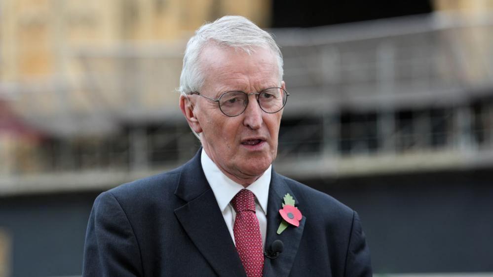 Hilary Benn has white hair and wears thin black glasses. He is wearing a black suit with a red tie. He has a red poppy on the left side of his blazer.