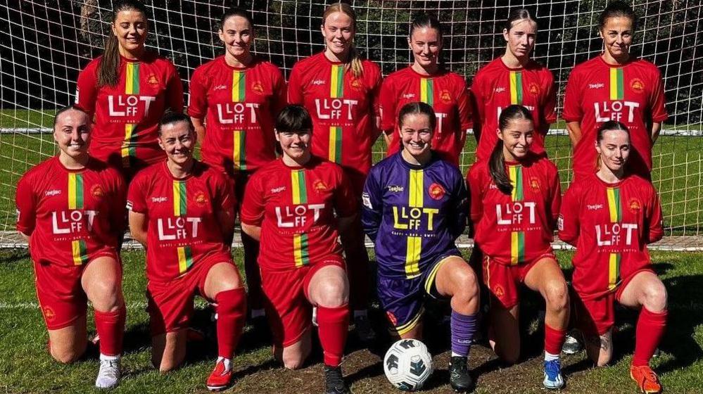 A group of young women from the CPD Merched Y Felinheli team, all dressed in red football kits - bar the goalkeeper, who is in blue - formally arranged in front of a goal.