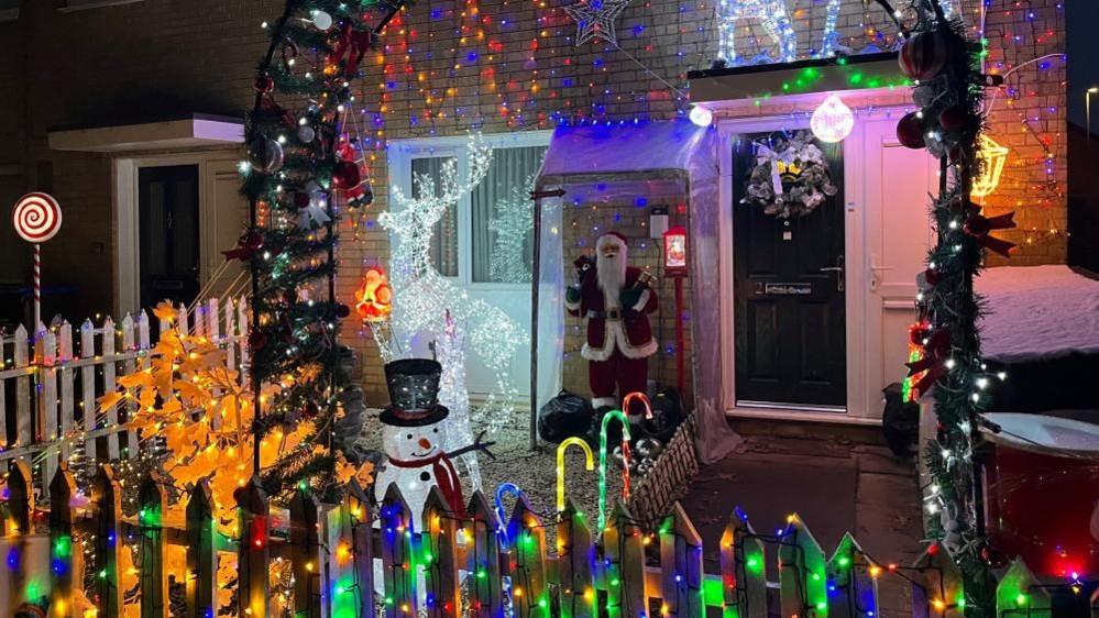 A wooden fence surrounded in multi-coloured Christmas lights. Behind it is a black metal arch decked out in more lights, baubles and Christmas tree material. A Santa stands outside the front door. 