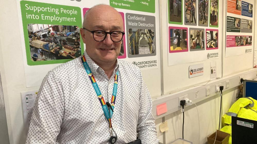 A man is wearing spotty shirt. He is standing in front of poster advertising people being supported into work.