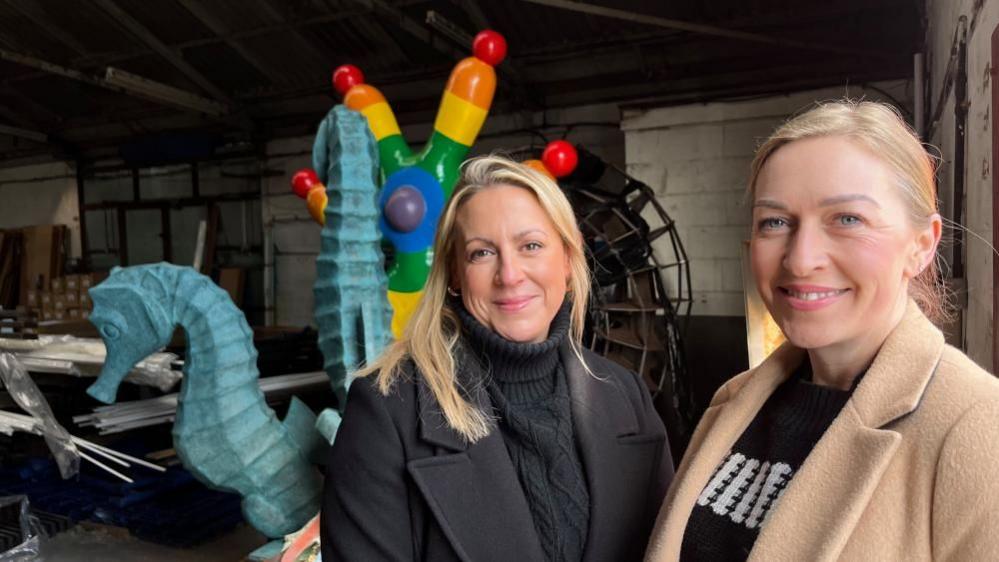 Natasha Hayes, centre, and Tracey Reed, right, stand inside a storage warehouse, with two sculptures behind them. Natasha is wearing a black polo neck jersey, with a black coat over, and Tracey wears a black patterned teeshirt, with a fawn coloured jacket. Both have blonde hair, with Natasha's flowing over her shoulders, and Tracey's hair tied back in a pony tail.