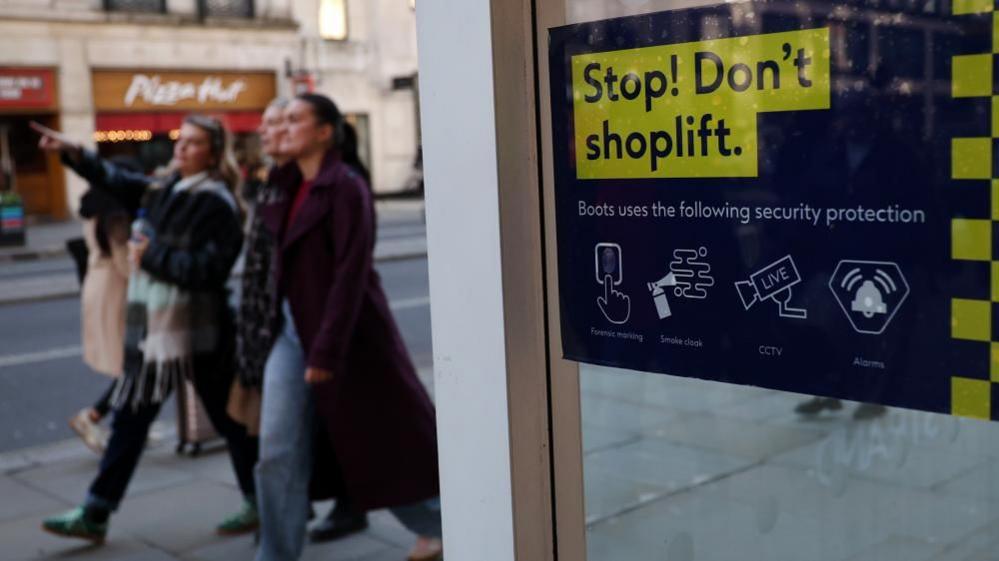A group of young woman walk along a street past a sign that reads "Stop! Don't Shoplift".