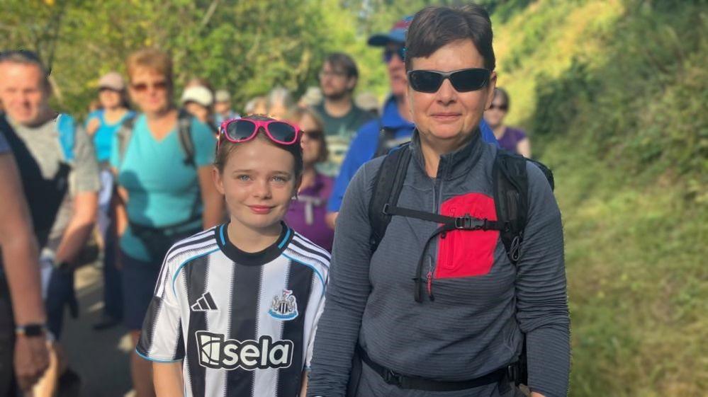 A young girl wearing a Newcastle United top and pink sunglasses on her head stands next to a woman wearing sunglasses, a grey walking top and a rucksack. They are outside on a footpath with hedges each side and among a crowd of people.