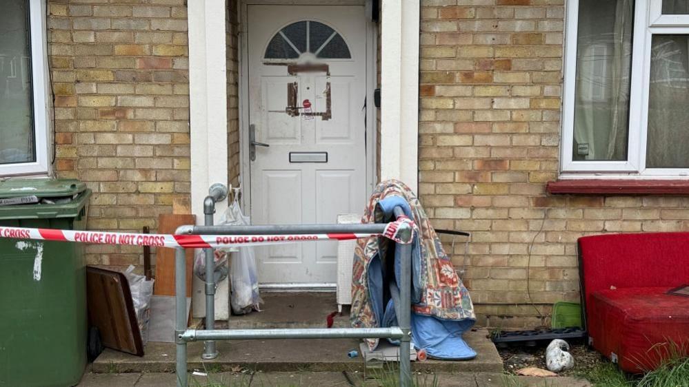 Police tape across a property in March. It shows metal railings, discarded items, a chair, bedding, outside the property and a green bin to the right. The front door is white, which has had a sign posted on it. 