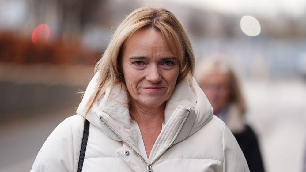 A woman with shoulder-length blonde hair looks at the camera. She wears a white puffa jacket. 