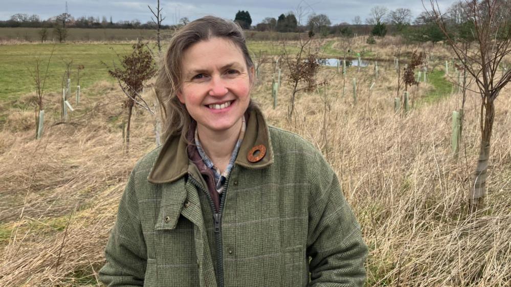 A woman stands smiling in a vast field with a lake and trees in the background. She is wearing a green tweed jacket. 