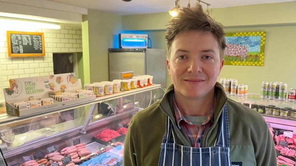 Tom Grant stands in his butchery shop. He is wearing a pink and blue checked shirt with a green fleece and a blue and white apron. He has slightly long hair which is combed up. He is standing before his refrigerated displays packed with meat, with other ingredients on the counter tops. On the walls is a price list, a picture showing the cuts of pork on a pig and a blue-light fly trap.