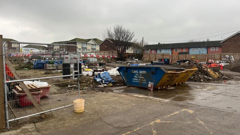 A building site with a pair of blue and yellow skips, and other building yard detritus and temporary fencing visible. The area is a concreted hardstanding of what was warehousing and commercial premises. Flats and maisonettes can be seen in the distance.