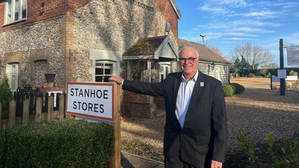 Terry Stork is wearing a black suit and white shirt. He is leaning on a sign which says "Stanhoe Stores" in front of a building which is clad with lots of round stones
