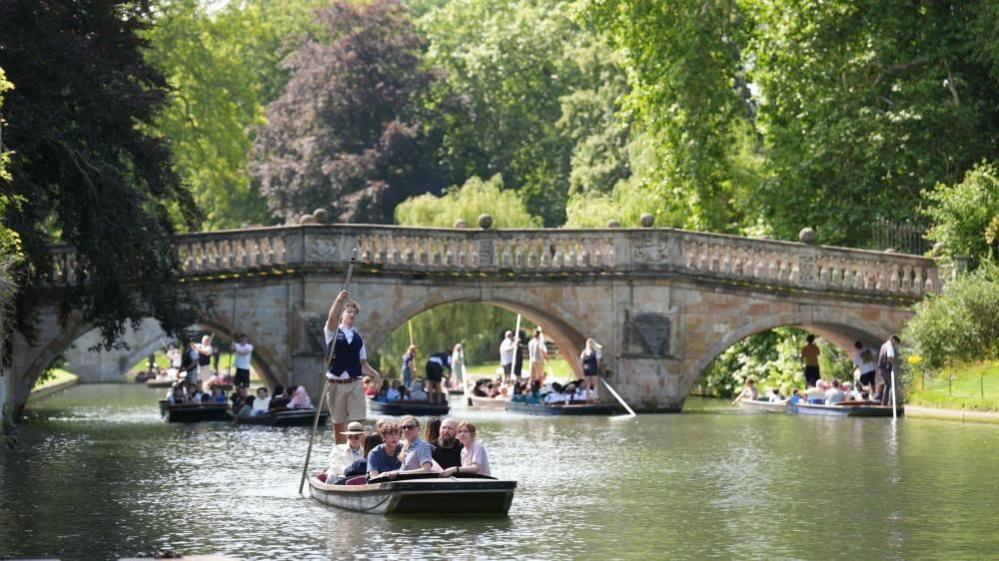 Four punts filled with people making their way along a river. A stone bridge is in the background. A tree overhangs on the left. A grass bank and shrubs are on the right.