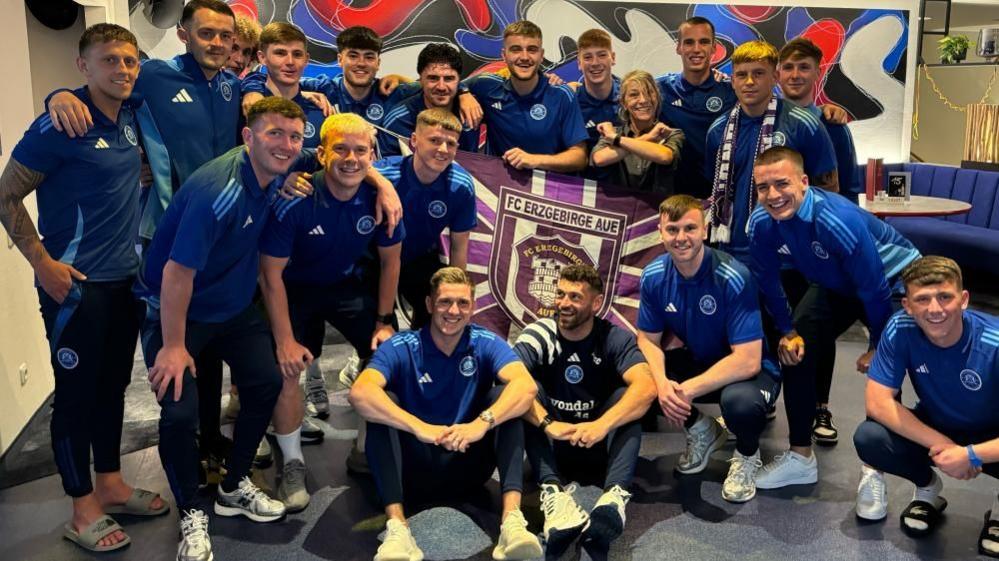 Glenavon players are arm in arm and smiling. They are wearing navy and blue polo shirts and are holding a purple and white Erzgebirge Aue flag.