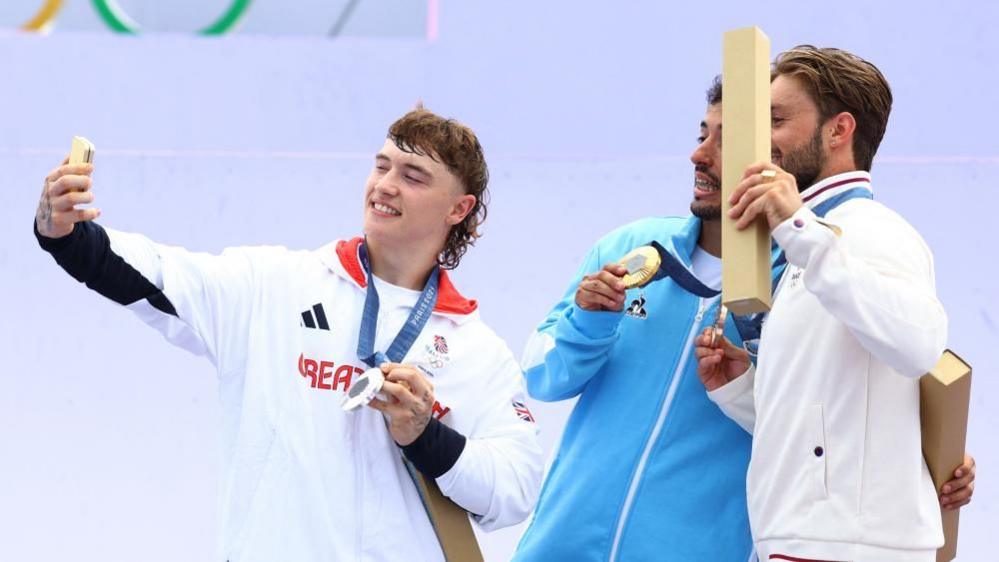 Kieran Reilly with dark brown mullet holds a smart phone with two men with dark hair and beards - all holding medals