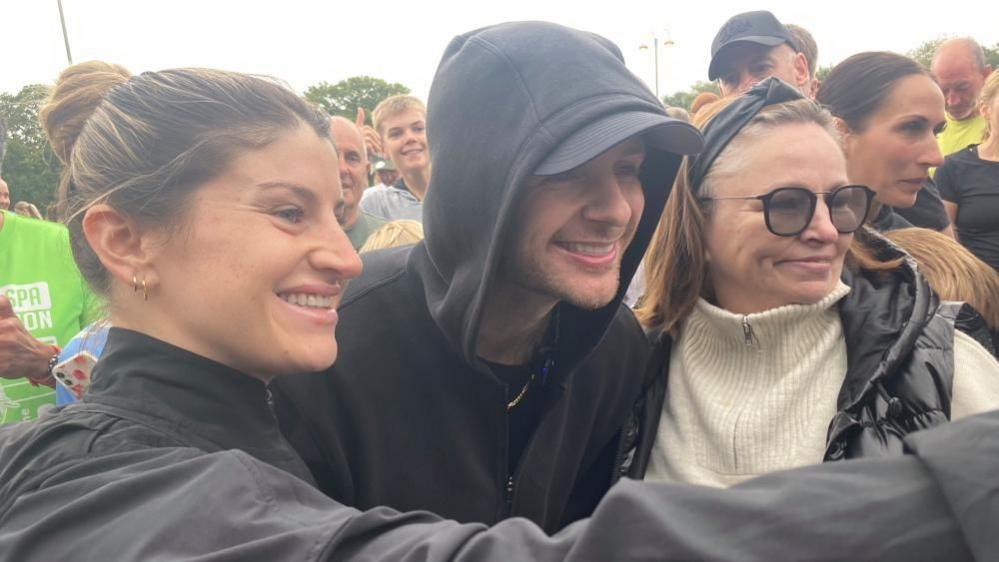 Two women who are taking a selfie with a man, singer Tom Grennan, who is wearing a black hoodie and has the hood up. They are outside before a run. 