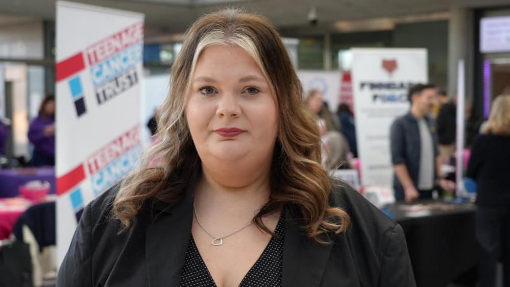 Kate Doe, from Community Action Norfolk,  is wearing a black dress and black top, with silver necklace. She has long auburn hair with a light streak of colouring at the front. She is standing near cancer charity stalls at an event at The Forum in Norwich.