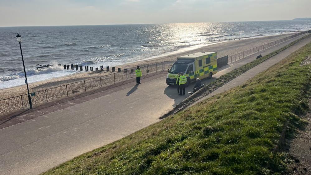 The sun shimmers on the sea at the south end of Gorleston beach. On the promenade is an ambulance and emergency service workers, including police and paramedics. 