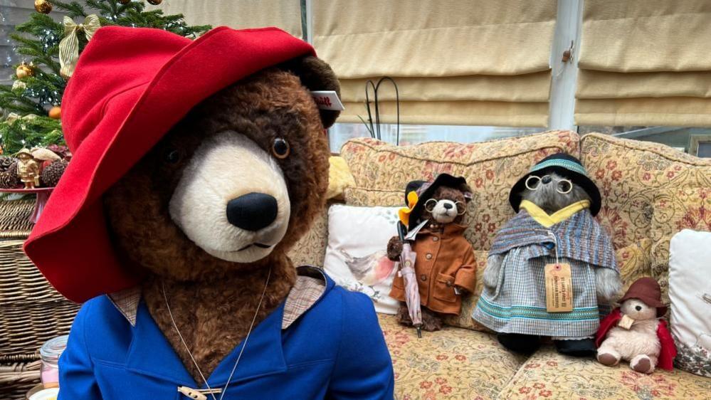 A closeup of a bear wearing a red hat and a royal blue coat. Behind it are three bears positioned on a sofa. 