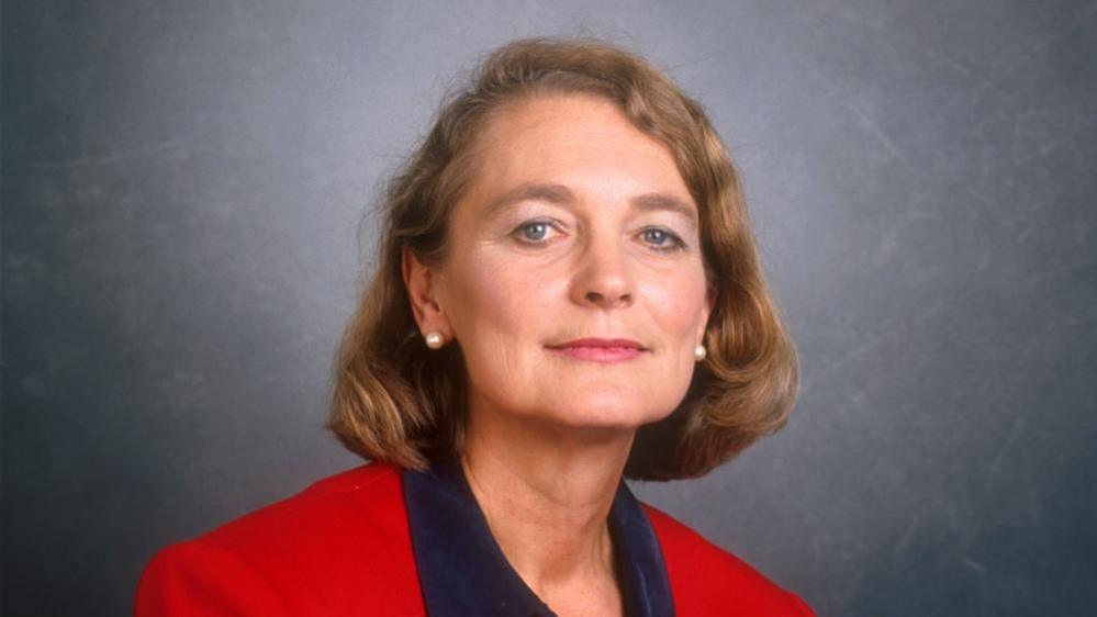 A House of Commons photograph of Anne Campbell against a grey background. She has shoulder-length blonde hair and pearl earrings. She is wearing a red jacket and a dark blue blouse.