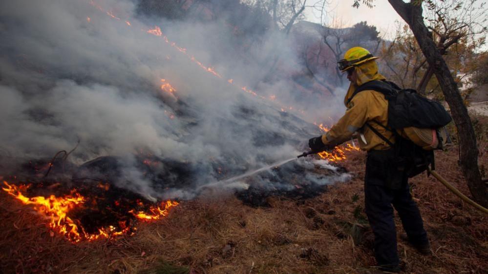 Firefighter with hose directed at fire on scorched ground with water pouring onto flames