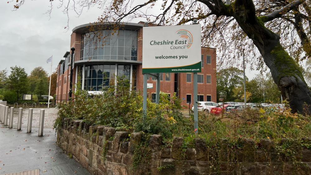 A red brick council building. There is a sign outside the building that reads "Cheshire East Council welcomes you". The sign is planted into a bed of shrubs with a stone wall in front. The council building has a circular entrance which is made up of glass. The red brick makes up the rest of the building's exterior. 