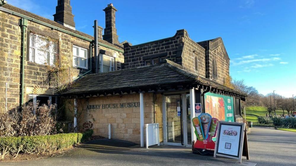 A brick building with a Historic England sign and ice cream sign outside. It has glass double doors and on the side reads Abbey House Museum in green lettering. 