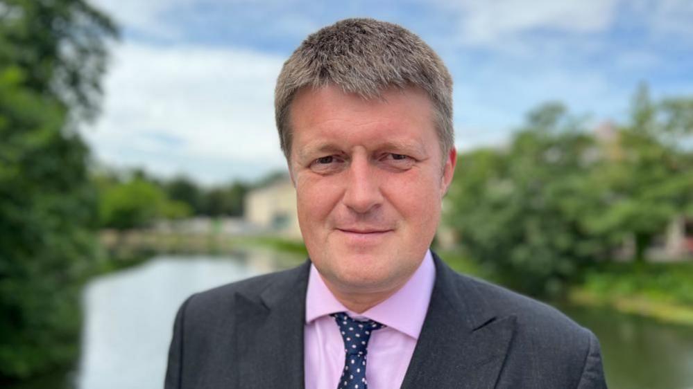 Wiltshire Council leader Richard Clewer looking direct to camera. He is wearing a suit and tie and stood in front of a blurred river.