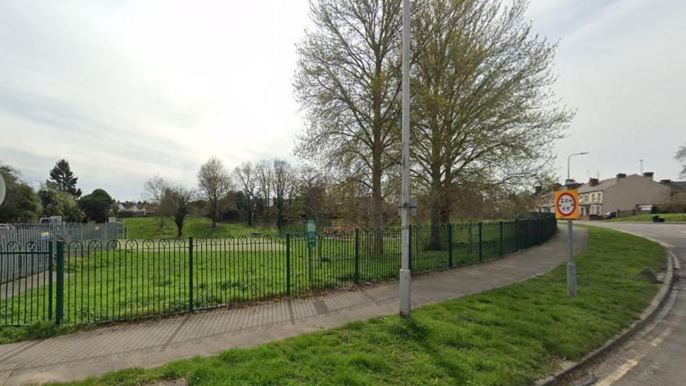 Metal fencing along the edge of a park with trees and homes seen along the edge.