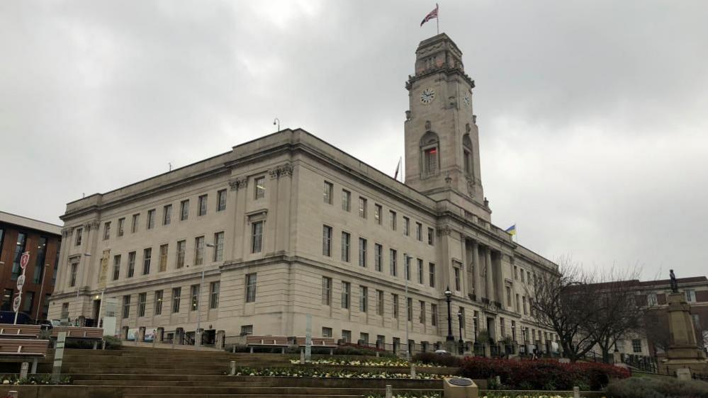 Barnsley Town Hall. It is a white stone building with four floors, and lots of windows around the building. It has a clock tower with a Union flag flying at the top.