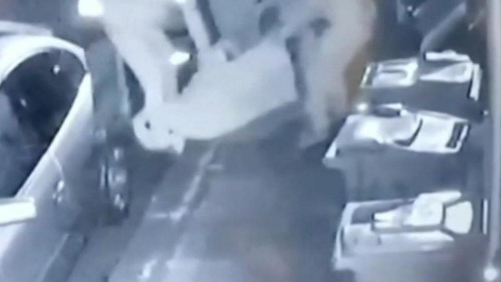 Black and white CCTV of a bundle being moved. There are two figures in the image. along with a car and several wheelie bins