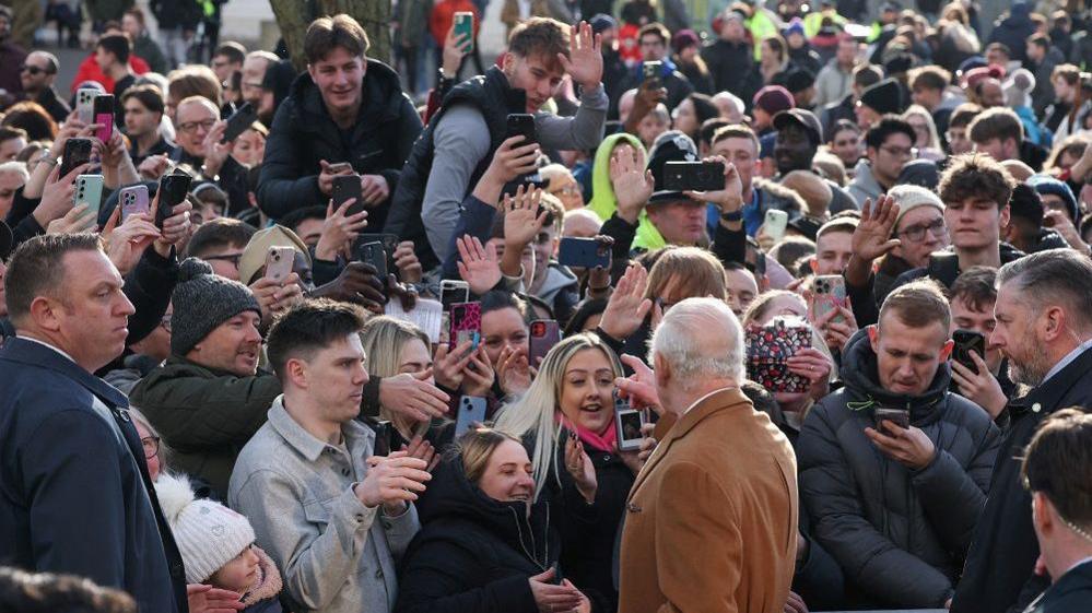 King Charles standing in front of a crowd of people. He's pointing and hands and phones can be seen waving back at him. 