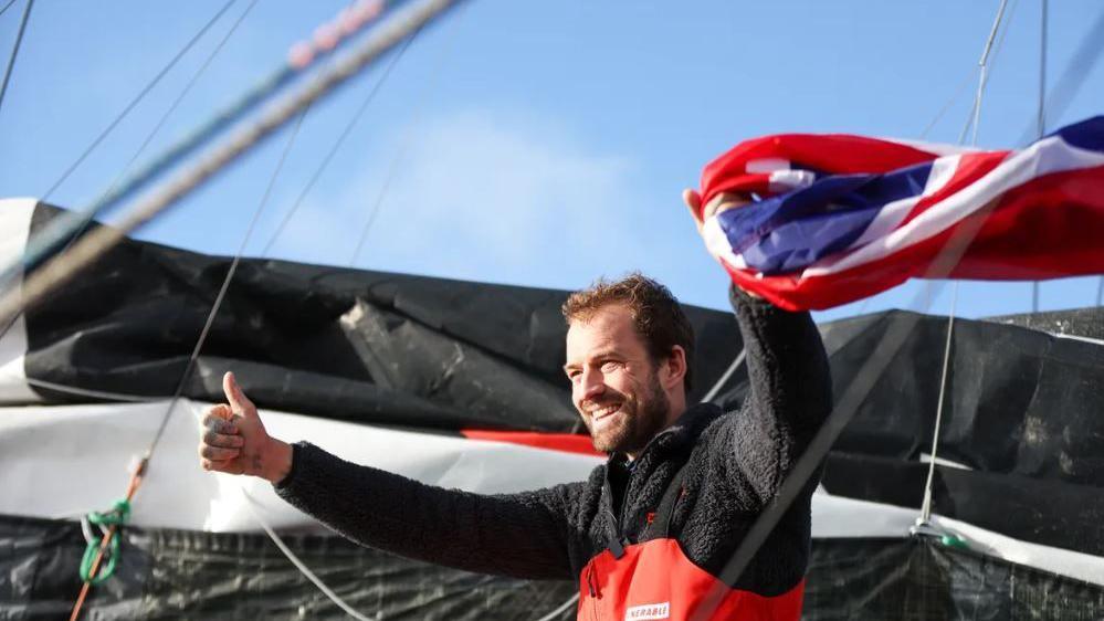 A man with a beard is smiling. He holds a union jack with one hand and puts a thumbs up with the other. He is wearing a thick top and sailing suit.  