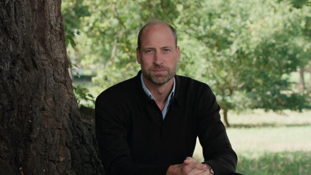 Prince William sits at the foot of a tree looking out beyond the shot of the camera.