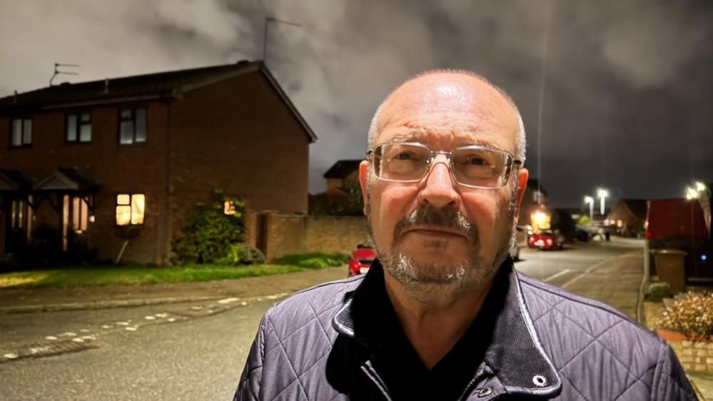 Graham Plant, wearing glasses and sporting a beard and moustache, which is greying. He is wearing a black quilted jacket, and is standing under a street lamp in the evening on a housing estate.