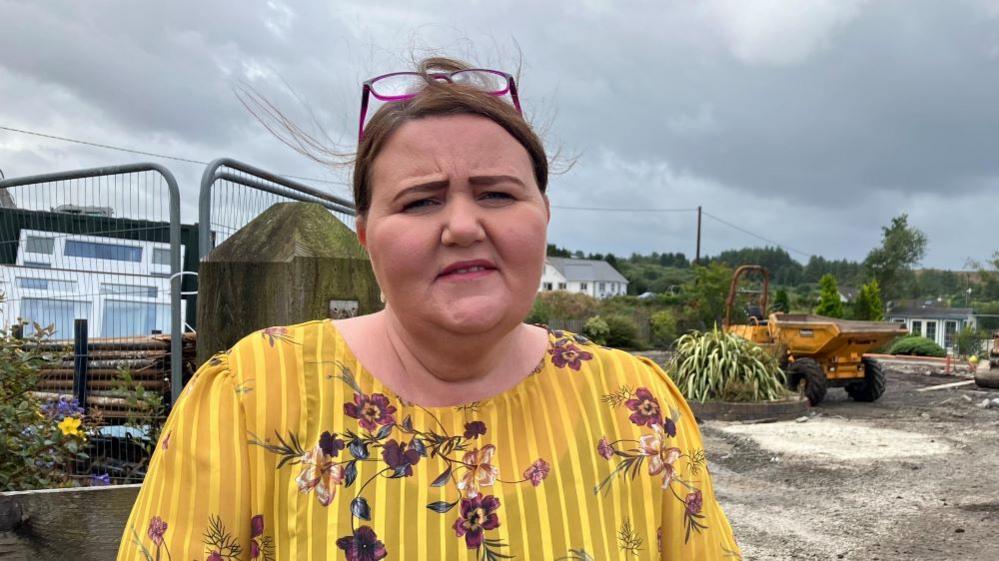 Joy Beard, wearing yellow top with floral design and purple glasses raised on top of her head, stands in front of a building site with diggers and barriers where her home once stood