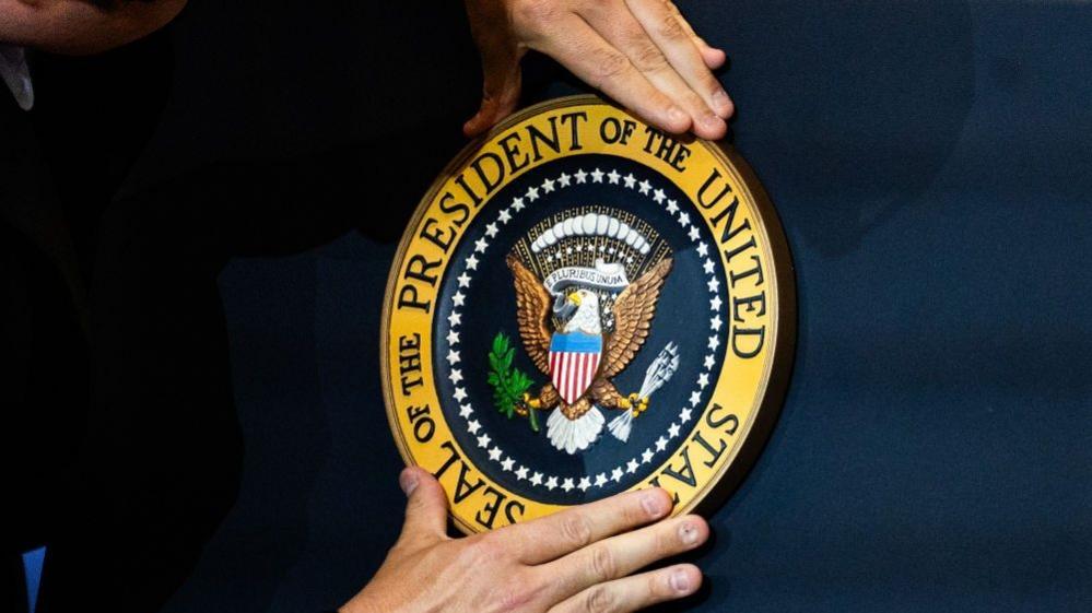 Two hands fix the golden US presidential seal on the front of a navy blue podium. 