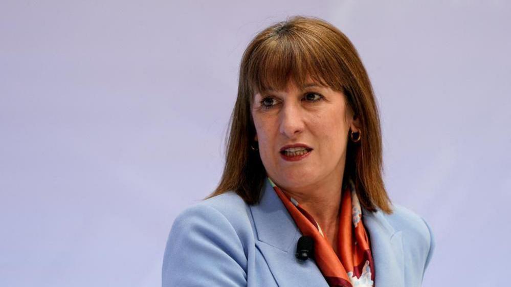 Headshot of UK Chancellor Rachel Reeves sitting with a plain purple background behind her. She has short, brown hair with a fringe, and is wearing a light blue jacket.