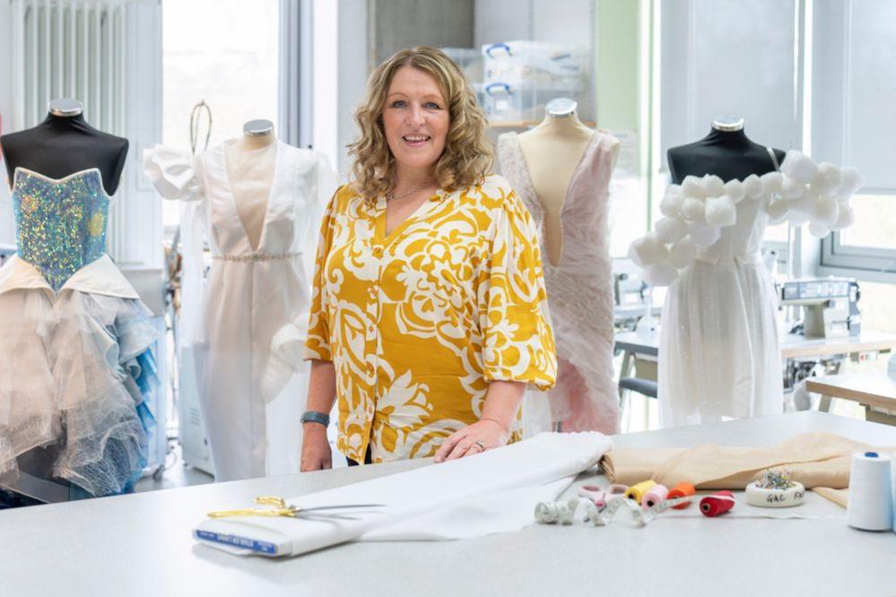 A woman with a yellow and white shirt and blonde hair stands in front of mannequins with four dress designs tacked onto them. It is a workshop setting.