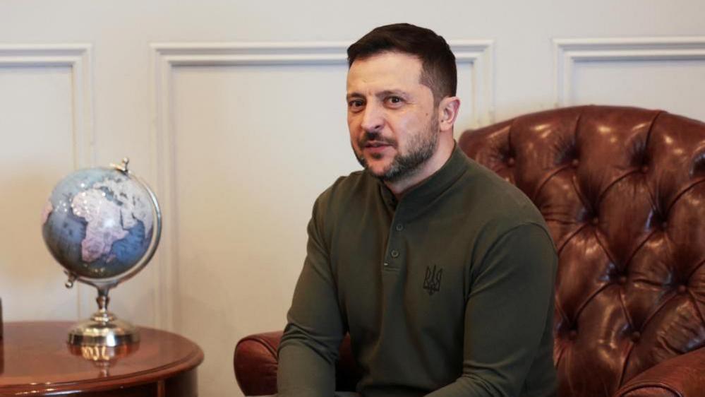  Ukrainian President Volodymyr Zelensky at Shannon Airport, 27 February - he is wearing a green jumper with a Ukraine crest faintly visible - he is sitting in a red leather armchair, with a globe of the world visible on a side table 