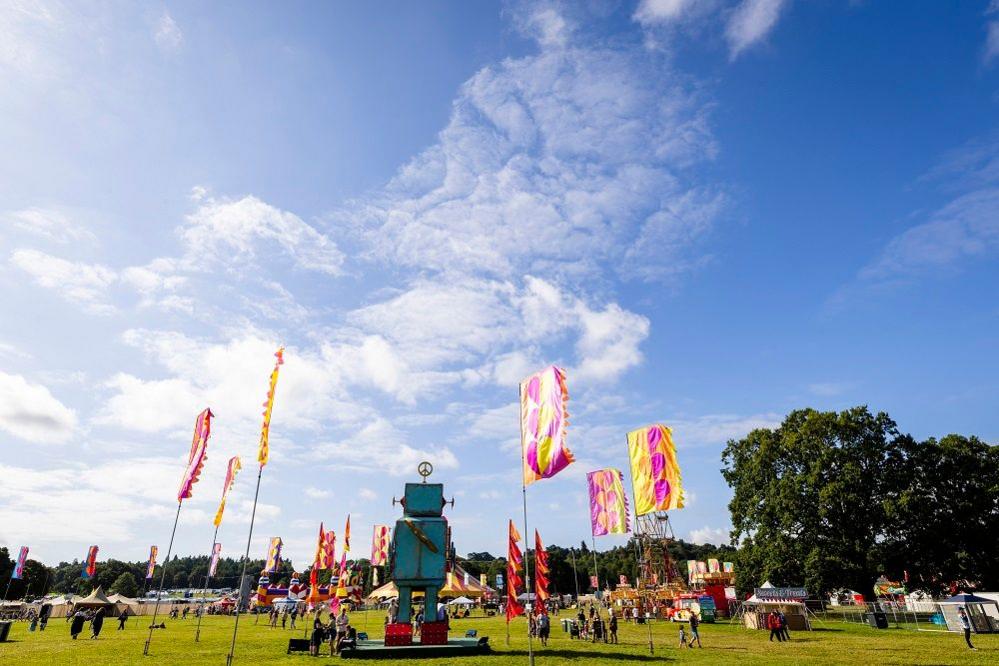 The festival under a bright blue sky with some whispy white clouds in the background. Two trees are alongside several flag poles with yellow and pink designs