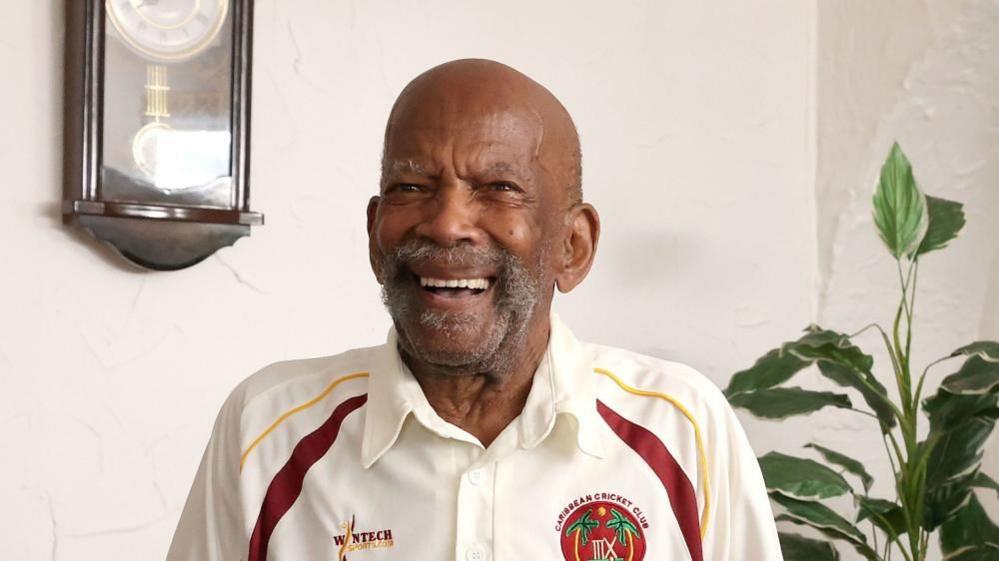 Alford Gardner wearing a Caribbean Cricket Club shirt with red and yellow details smiles at the camera.