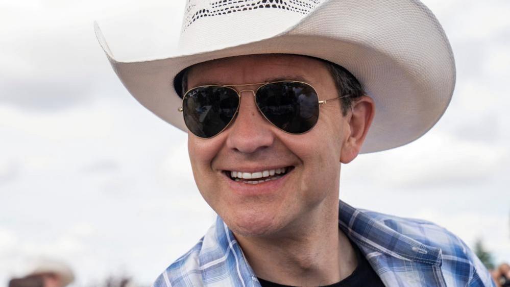 Pierre Poilievre sports a wide grin along with aviator sunglasses, a white cowboy hat, and a blue and white plaid shirt over a black T-shirt.
