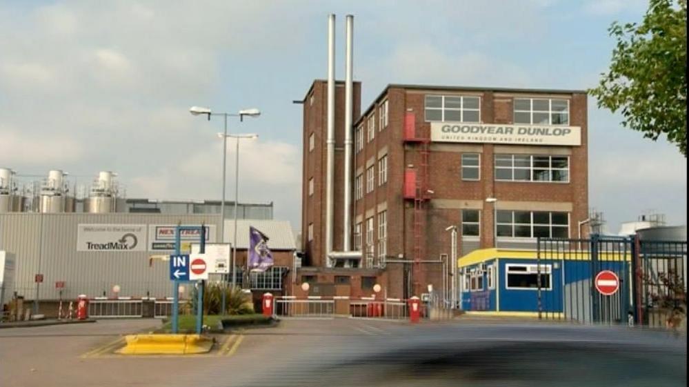 A brown brick industrial building with a flat room and large silver chimney pipes protruding from the side. It has a sign that says Goodyear Dunlop. In the background a metal clad industrial building is visible