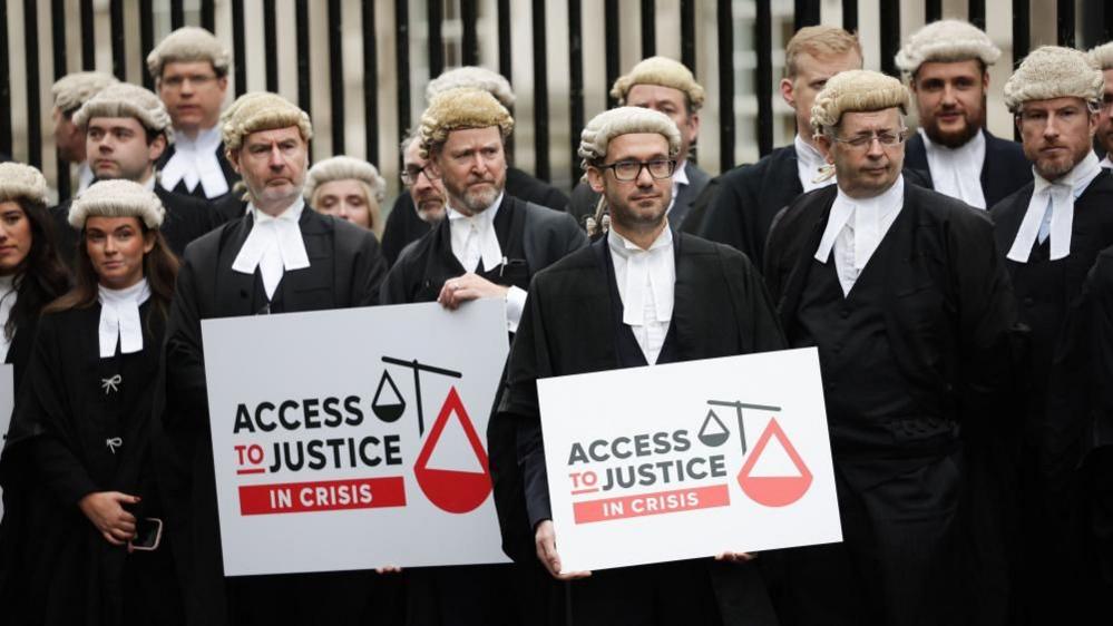 A crowd of barristers wearing white wigs, black gowns and white collars stand at a picket line outside Belfast Laganside court. They are holding signs that say access to justice in crisis.