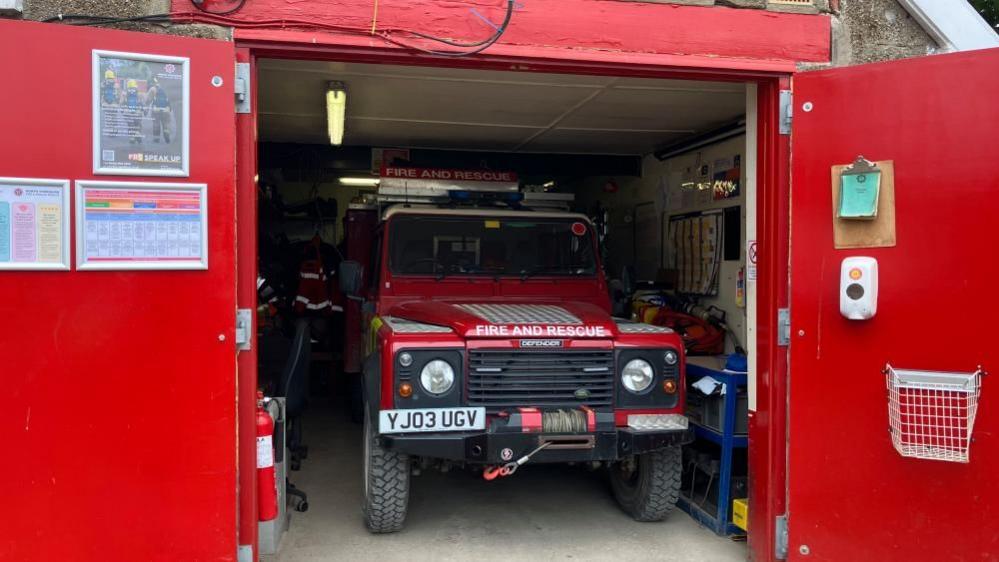 A Land Rover vehicle in a shed, inches away from equipment racks on one side, with about a foot width on the other side, giving access to get into the vehicle but not much more. Posters are on the inside of the red doors, which are open.