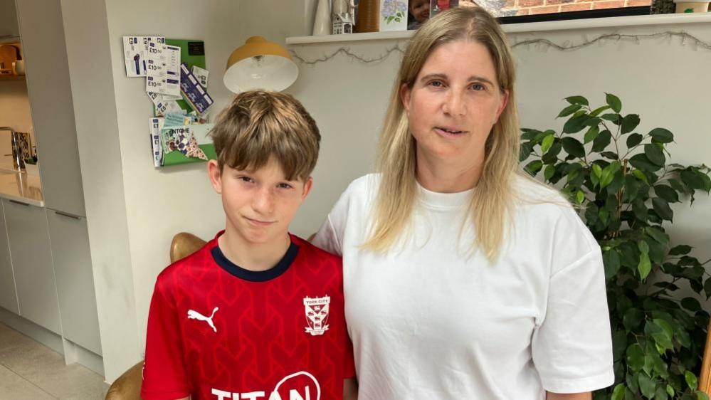 A boy and his mum look into the camera, in a room with white walls and a plant behind them. There is also a noticeboard with coupons on it, and fairy lights. He is wearing a York City FC football shirt, and she is wearing a white T-shirt.