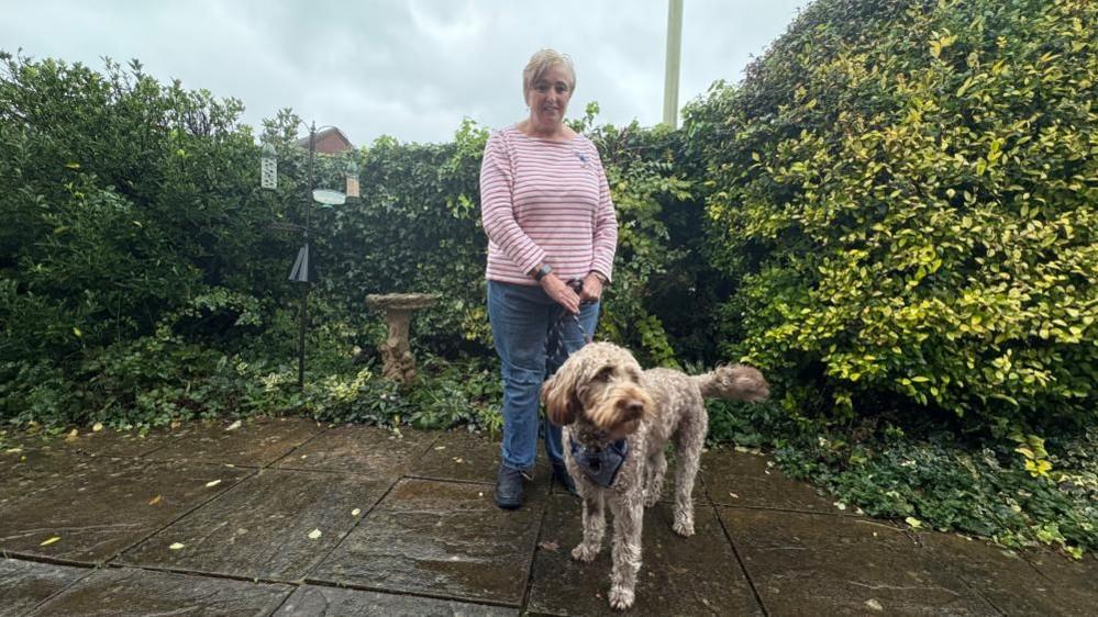 A woman with short blond hair and a red and white stripey top and jeans stands on a patio with a hedgerow behind her. She holds a caramel coloured Labradoodle dog on a lead.