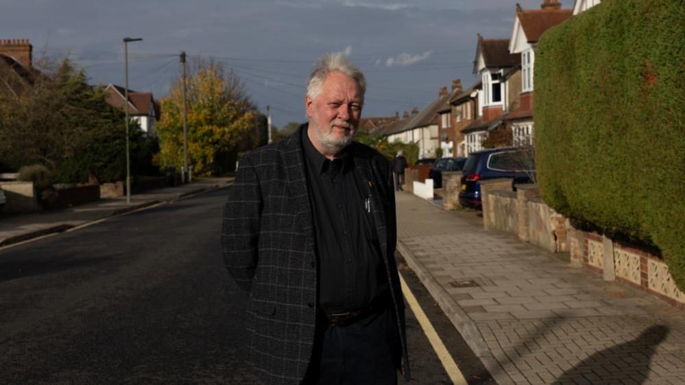 Mr Kent stands in the road with his hands behind his back wearing a black suit with a black shirt. He has grey hair and a short grey beard. There is a green hedge to the right of him.