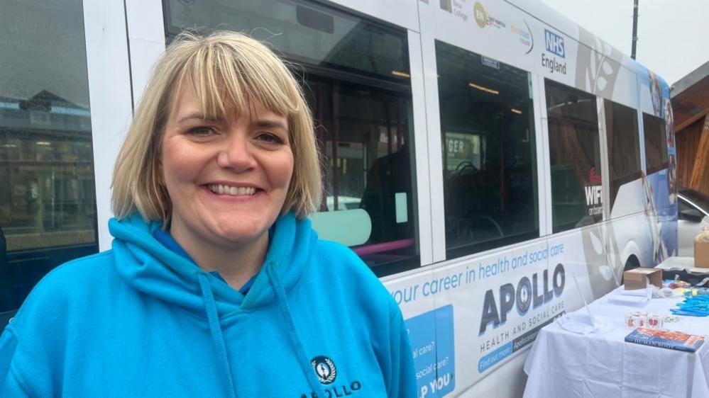 Sonia Loveday, with blonde hair and wearing a light blue hooded top, standing by the Apollo Health and Social Care roadshow bus