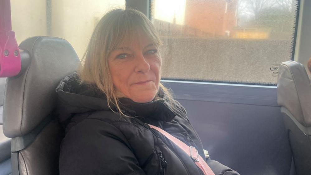 Toni Norman has long blonde hair, is smiling and is wearing a black jacket with a pink bag strap across her chest. She's sitting on a grey leather bus seat and there's a pink grab pole in the upper left of the image. The window of the bus is dirty and it is not possible to decipher what is in the background. 