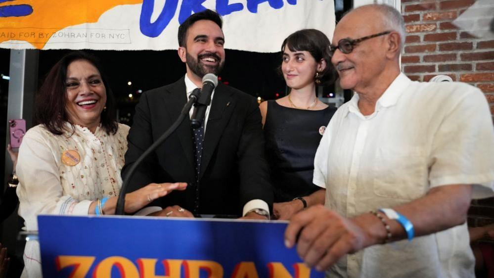 Zohran Mamdani stands at a lectern next to his parents Mahmood Mamdani and Mira Nair, and his wife Rama Duwaji, during a watch party for his primary election