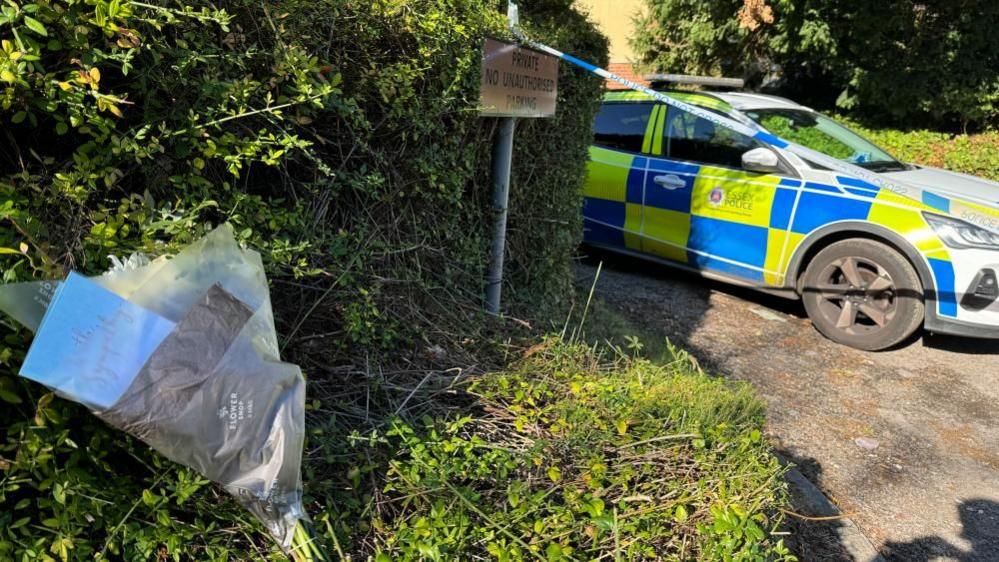 A bunch of flowers left by the side of a road, and a police car by police tape, a sign says Private No Unauthorised Parking